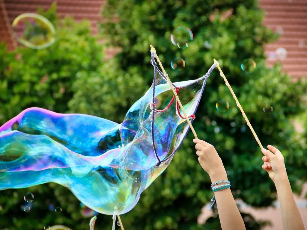 A huge bubble with rainbow colours being formed with a large bubble wand being held by a woman, outdoors with greenery in the background
